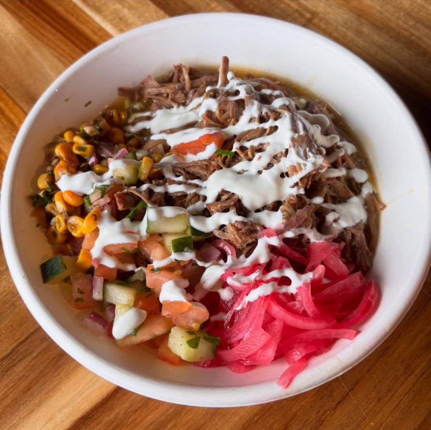 A Braised brisket bowl loaded with corn salsa, tomato cucumber salad and pickled onions